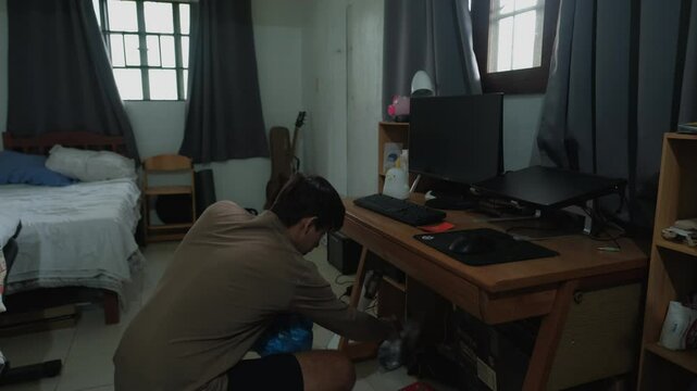 Young Caucasian man organizing and cleaning under messy desk in bedroom. Wide shot.