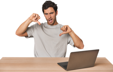 Hispanic young man working on laptop showing thumb down and expressing dislike.
