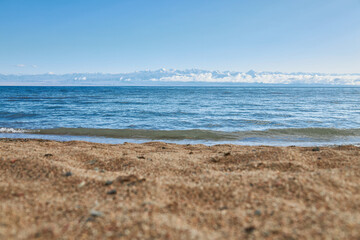 Sandy beach, coast of southern shore of big lake Issyk-Kul, summer vacation in Kyrgyzstan. Clear blue sky, emerald colored water of pond, small wave. Kirgiziya travel destination. Mountains in clouds