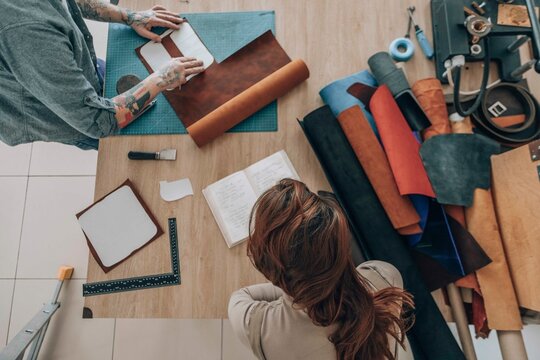 Businessman measuring leather patch near businesswoman reading diary at workbench