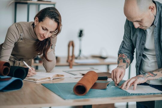 Businessman cutting leather patch near businesswoman writing in diary at workbench