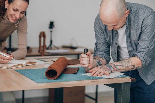 Smiling businessman cutting leather near businesswoman writing in diary at workbench