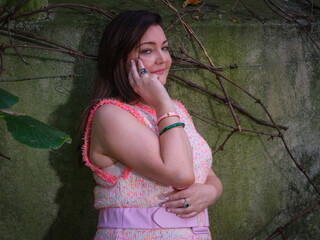 A woman in a pink sleeveless dress stands pensively against an aged, vine-covered green wall in a...