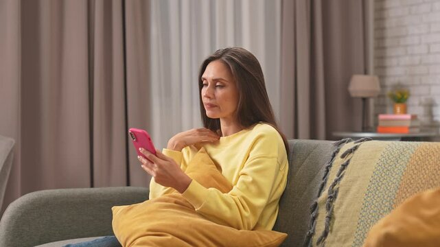 Young woman spending day at home sitting on the sofa in the living room, texting on smartphone and waiting for reply, worried sad impatient expression.