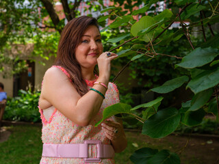A woman in a pink sleeveless dress with matching heels leans casually against an old, vine-covered stone wall in a garden setting.