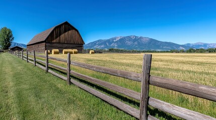 Panoramic landscape view of a wooden barn nestled in a field of golden hay, with a rustic wooden fence bordering the foreground. A mountain range