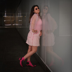 A woman in a pink dress and sunglasses leans against a reflective glass wall, smiling confidently...