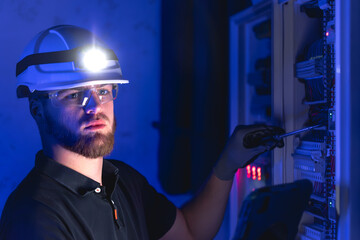 A male electrician works in a switchboard in overalls against the backdrop of emergency lighting.