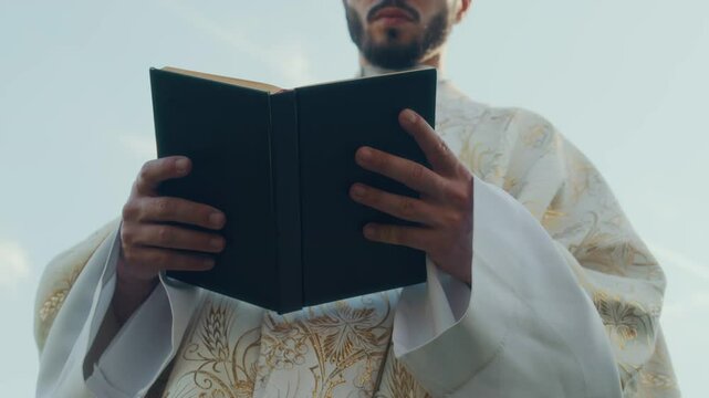 Midsection view from below of young priest dressed in golden white chasuble reading holy book outdoors