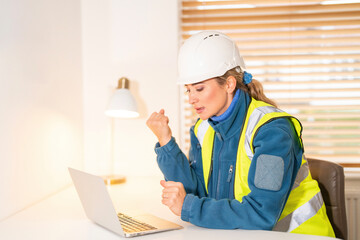 Female Engineer Construction worker focuses on laptop while wearing safety gear in bright office space
