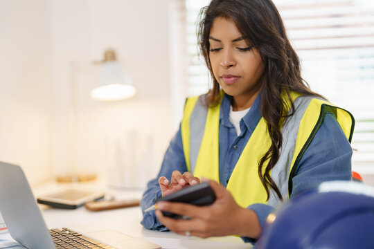 Female Construction worker engages with smartphone while sitting at desk in office wearing safety vest during daytime hours - Powered by Adobe