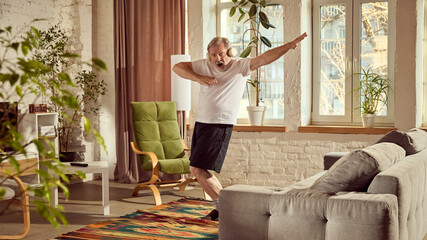 Elderly man in home wear listening to music in headphones and dancing in modern living room at home with potted plants and grey couch. Concept of happiness, elderly lifestyle, relaxation