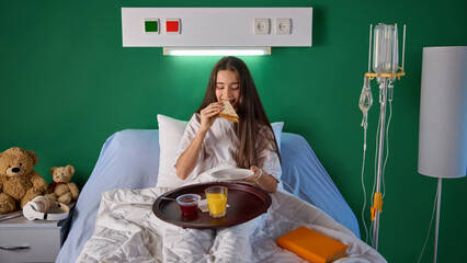 Teen girl sitting in bed in hospital ward and eating dinner served on food tray, sandwich and...