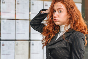 Confused woman with red hair in a professional outfit stands near bulletin board trying to find...