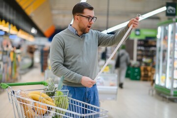 Portrait of an elegant man with shocked emotions holding very long shopping list while buying food in the supermarket