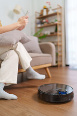 Asian woman using her smartphone while a robotic vacuum cleans the floor in a modern living room