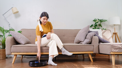 Asian woman turning on a robotic vacuum while sitting on a sofa in a cozy living room