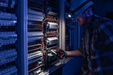 A male electrician works in a switchboard in overalls against the backdrop of emergency lighting.