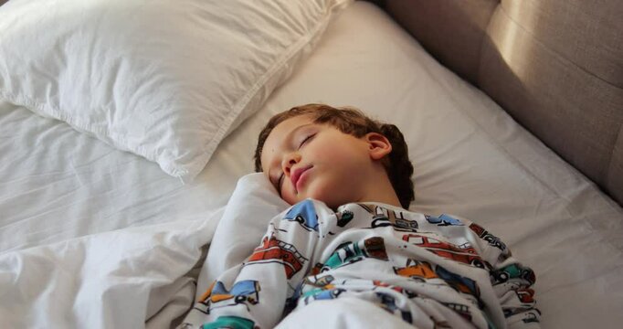 Innocent little boy enjoys dreams sleeping in bed with soft linen. Cute child in pajamas with cars enjoys magic dreams under soft morning sunlight