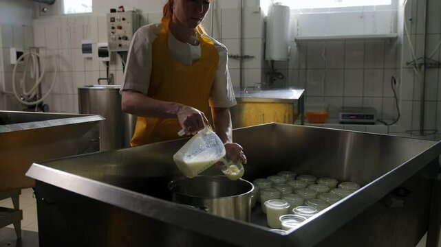 A female employee of a cheese factory stands in the workshop next to a cart and fills plastic bottles with milk. Kefir bottling at the factory, packaging of dairy products, food industry.