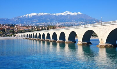 Fototapeta premium Arched bridge over turquoise water, snow-capped mountains, coastal town