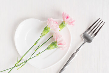Top view of ceramic heart shaped plate with cute pink carnation flowers with green stems served on white wooden table with silver fork prepared for celebration dinner of valentine holiday or date