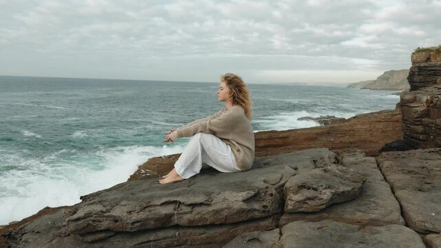 A thoughtful woman sits on a rocky cliff by the sea, gazing into the distance, reflecting on life with the ocean breeze around her.