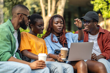 Young african american friends chilling with coffee on park bench, watching movies or video calling on laptop. Black students laughing together, gesturing and having outdoor fun, sharing conversation