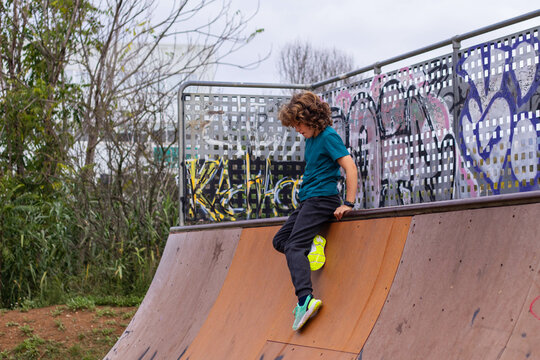 Boy at a skate park, sitting on the edge of a ramp with graffiti in the background. He is wearing a blue shirt, dark pants, and colorful shoes.