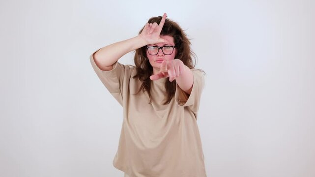 Brunette young plus size woman in beige t-shirt making loser gesture, L sign on forehead, teasing, accusing for defeat, expressing disrespect. Indoor studio shot isolated on white background. you lose