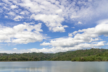 Landscape of a Calm Lake with Clear Water, Surrounded by Lush Green Forest Hills under a Bright Blue Sky Filled with White Fluffy Clouds, Showcasing Tranquil Tropical Nature Perfect for Travel