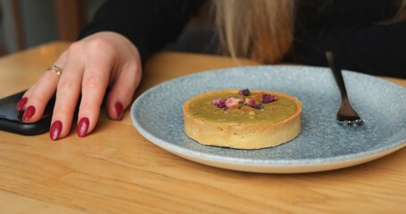 Pistachio tart on plate and anonymous woman hand with red nail polish on table. Unrecognizable female person in black long sleeve clothes sits at wooden table with delicious dessert on it. 4k footage.