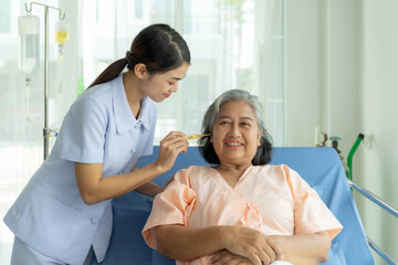 Obraz premium Asian nurse wearing a light purple uniform attentively taking care of an elderly Asian woman inhospital gown, measuring her temperature with a thermometer while smiling warmly in a clean hospital room