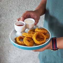 A person holds a plate of golden onion rings accompanied by two small cups of dipping sauces, ready to enjoy in a relaxed setting