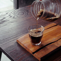 Barista pours hot coffee from a glass carafe into a glass on a wooden cutting board at a cozy cafe...