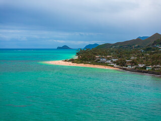 Turquoise ocean meets sandy beach lined with palm trees and homes, with lush hills and a small...