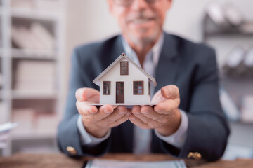 Real estate agent showing a scale model house in his office, with paperwork on the wooden desk and shelves in the background, offering property for sale or rent