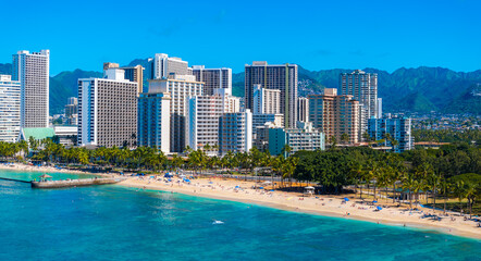 Obraz premium Turquoise ocean waters meet sandy shores and palm trees at Waikiki Beach, with Honolulu's skyline and the Ko olau mountains in the background.