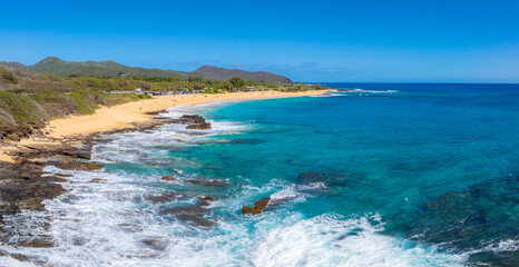 Aerial view of a beach on Oahu island, Hawaii, featuring golden sand, turquoise waters, rocky outcrops, green hills, and a small road with parked vehicles.