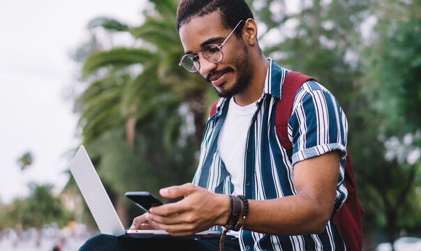 Young millennial dark skinned guy holding mobile phone reading information about online booking, hipster guys spending time time outdoors holding mobile phone working via laptop on freelance