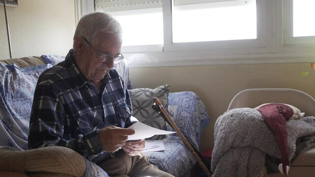 older man on sofa reviewing paper invoice, blue checked shirt and protective fabric armchair, in the background leaning on canes
