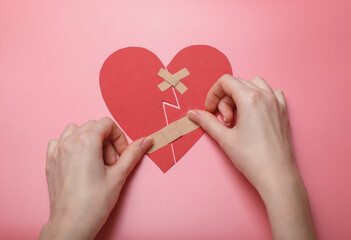 Hands carefully applying band-aids to broken red paper heart on pink background, symbolizing healing, love recovery, emotional repair. Touching concept for heartbreak, forgiveness, relationships, top 