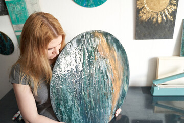 Young caucasian female artist with long hair examining a round abstract painting in studio setting.