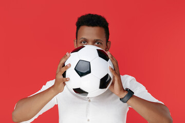 Handsome african american young man holding soccer ball in front of face over red background