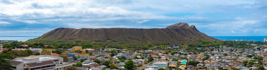 Obraz premium Diamond Head Crater rises above a residential area on Oahu, Hawaii, with rugged slopes, the Pacific Ocean in the background, and partly cloudy skies.