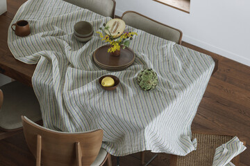 Simple organisation of dining table with ceramic dinnerware, linen tablecloth, dining chairs, spring flowers, lemons, melon, artichoke. Home decoration concept