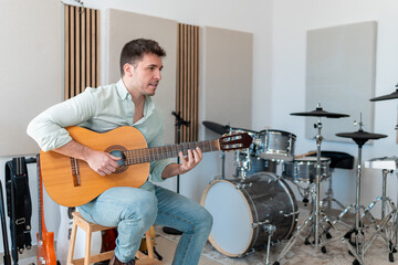 Fototapeta premium A young man playing an acoustic guitar in a professional recording studio with drums and soundproof panels in the background