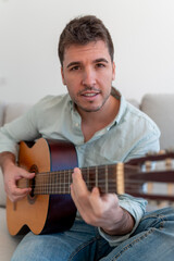 A guitarist sitting on a sofa, playing an acoustic guitar while looking at the camera with a...