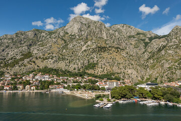 Kotor, Montenegro - July 2, 2024: Landscape, new and historic town with its ramparts and moat in front of tall mountain range under blue cloudscape