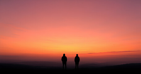 Fototapeta premium Two figures stand silhouetted against a vibrant sunrise. 